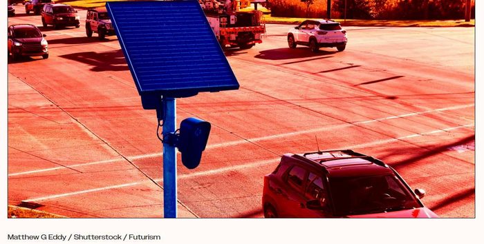 A Flock automatic license plate reader camera mounted on a pole with a solar panel at a suburban intersection.
